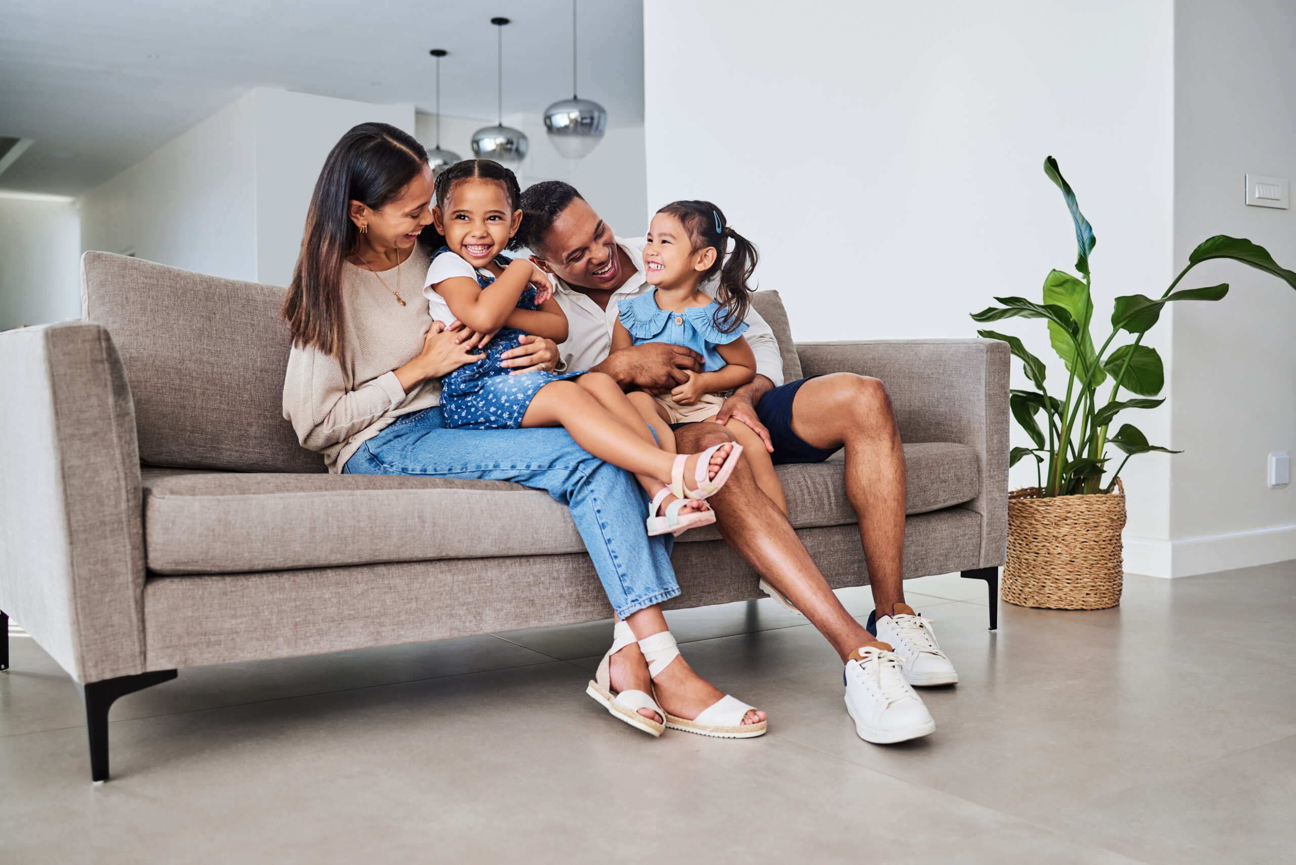 Family, children and laugh with a girl, sister and parents sitting on a sofa in the home living room together. Kids, laugh and humor with a mother, father and daughter siblings bonding in a house.