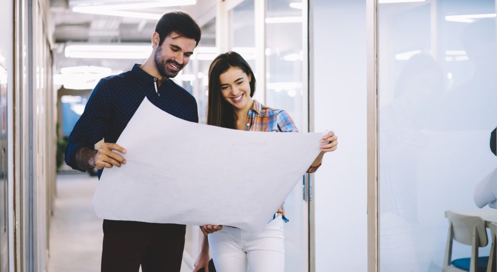 Happy young colleagues in casual wear watching blueprint of new architectural project and looking satisfied with final result in office hallway
