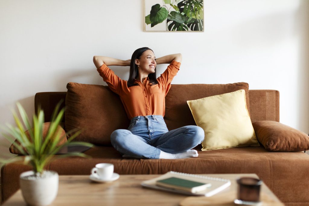 Contented relaxed woman with her hands behind head, reveling in serene and joyful break on comfy sofa in well-lit, stylish living room, free space